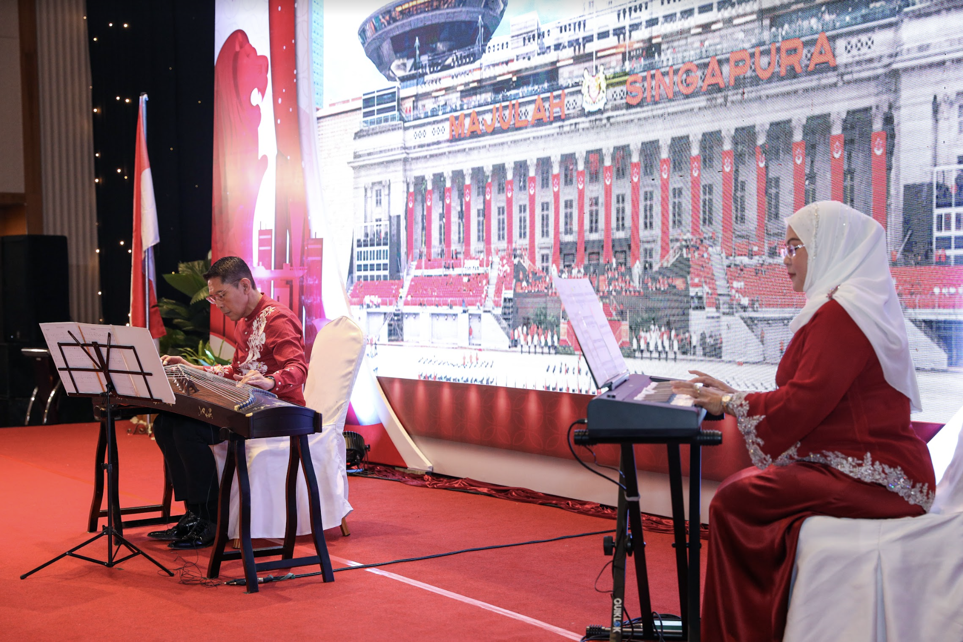 Two musicians, one man and one woman in a hijab, play instruments before a "SINGAPURA" backdrop.